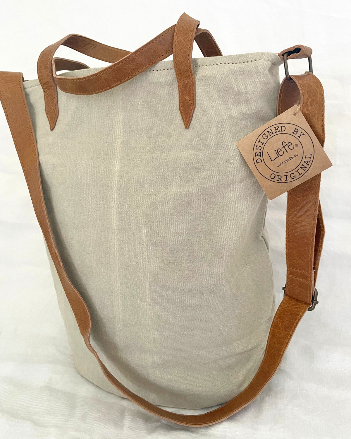 A canvas tote bag in a light beige color with brown leather handles and a matching adjustable shoulder strap. A paper hangtag on the side reads 'DESIGNED BY etsy ORIGINAL'.