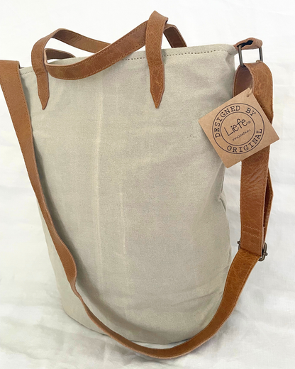 A canvas tote bag in a light beige color with brown leather handles and a matching adjustable shoulder strap. A paper hangtag on the side reads 'DESIGNED BY etsy ORIGINAL'.