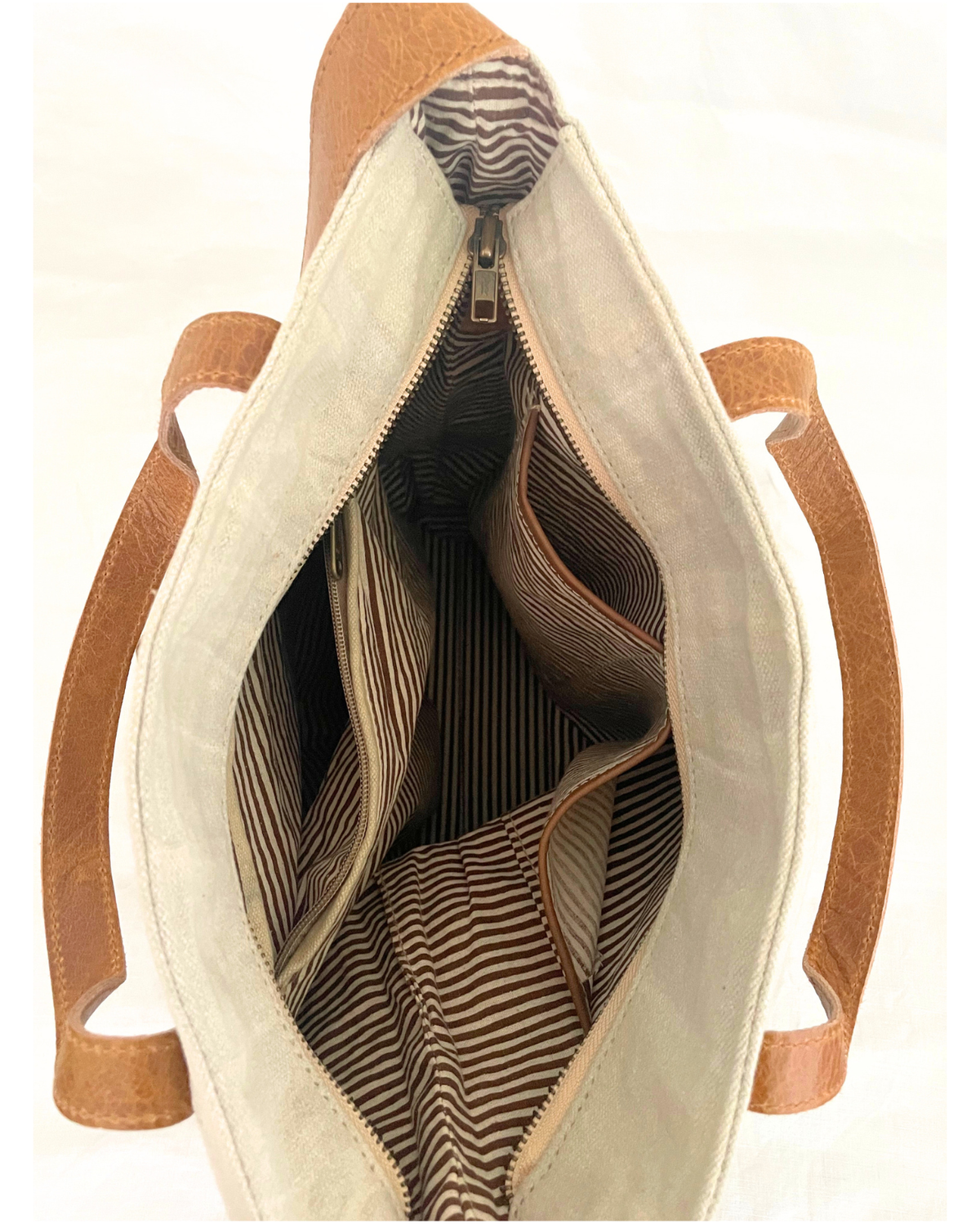 Open view of a tote bag showing the interior with cream canvas lining, brown leather handles, and a striped fabric lining inside.