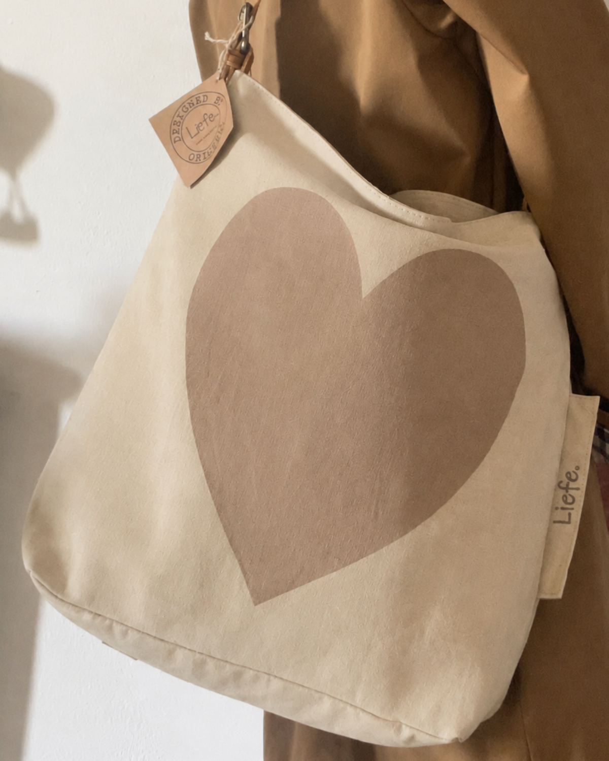 A canvas shoulder bag in warm off-white with a large muted tan heart printed on the front, hanging against a brown garment. A small leather tag reads 'LIEFE' and a side fabric tag also shows 'LIEFE'.