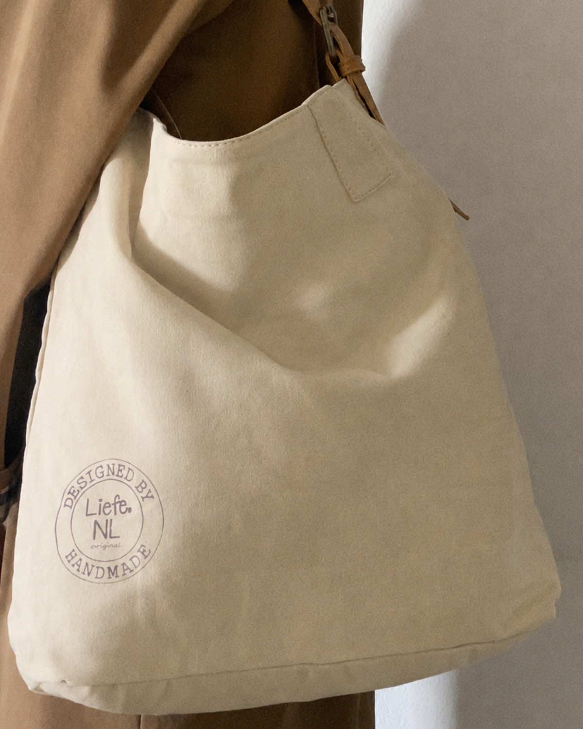 A beige canvas shoulder bag with a brown leather strap, featuring a circular 'Designed by Liets NL Handmade' stamp on the front, shown worn over the shoulder of a person in a brown jacket.