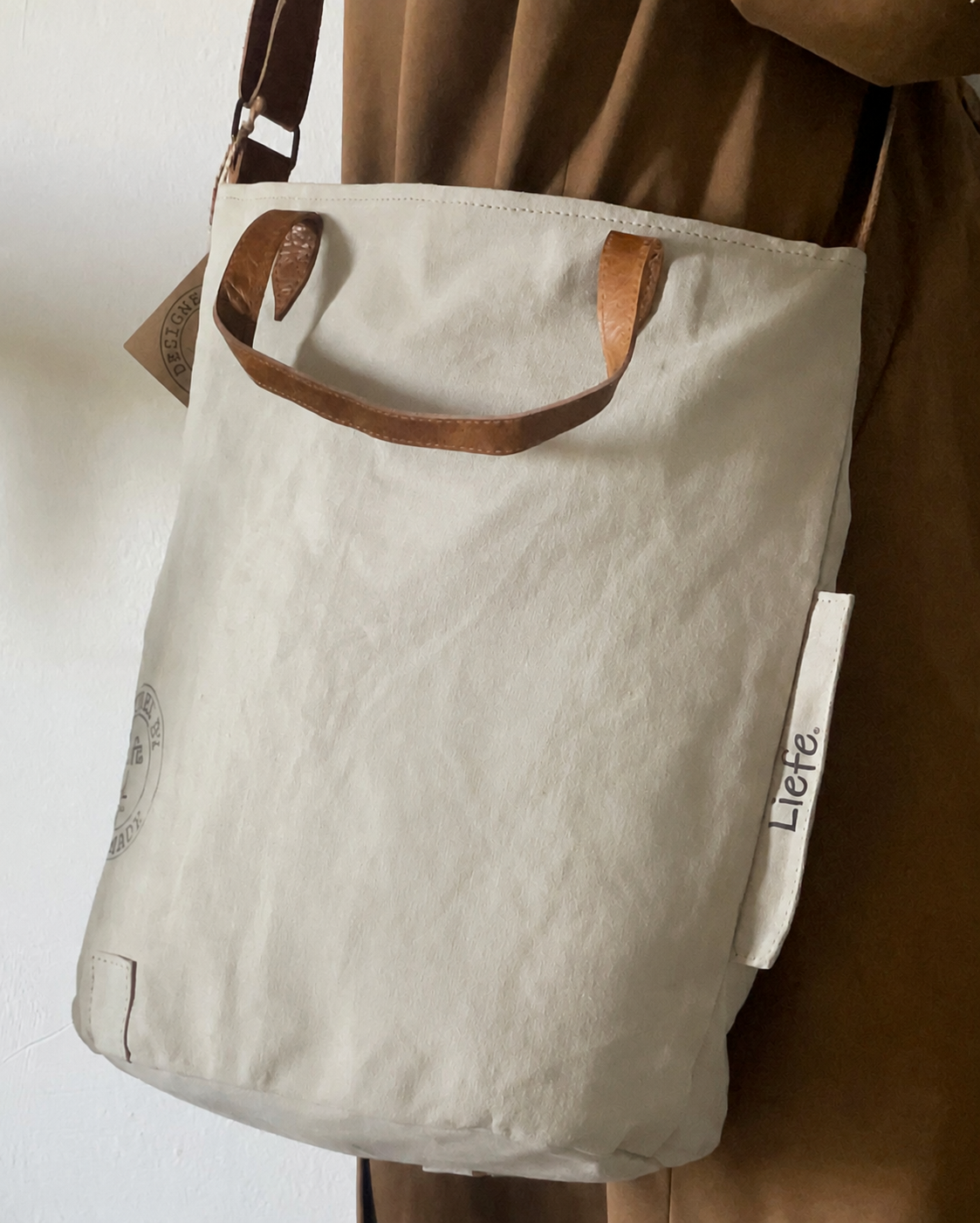 A cream canvas tote bag with brown leather handles and a brown shoulder strap, worn by a person in a brown dress. The brand name 'Liete' is visible on a white fabric tag hanging from the side.