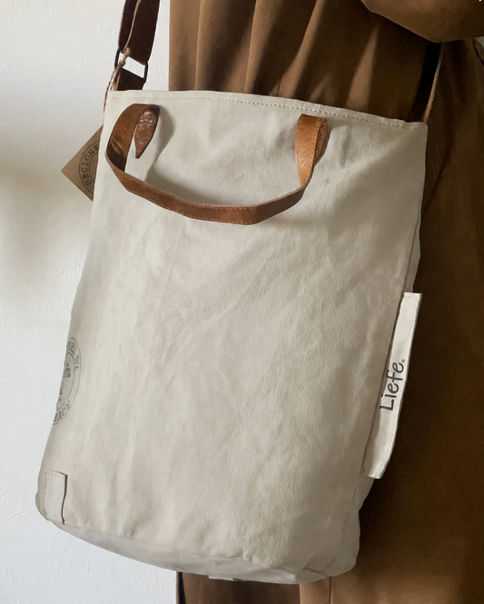 A light beige canvas tote bag with brown leather handles, a matching adjustable shoulder strap, and a hanging leather brand tag that reads 'DESIGNED BY LIFE TOUGH'.