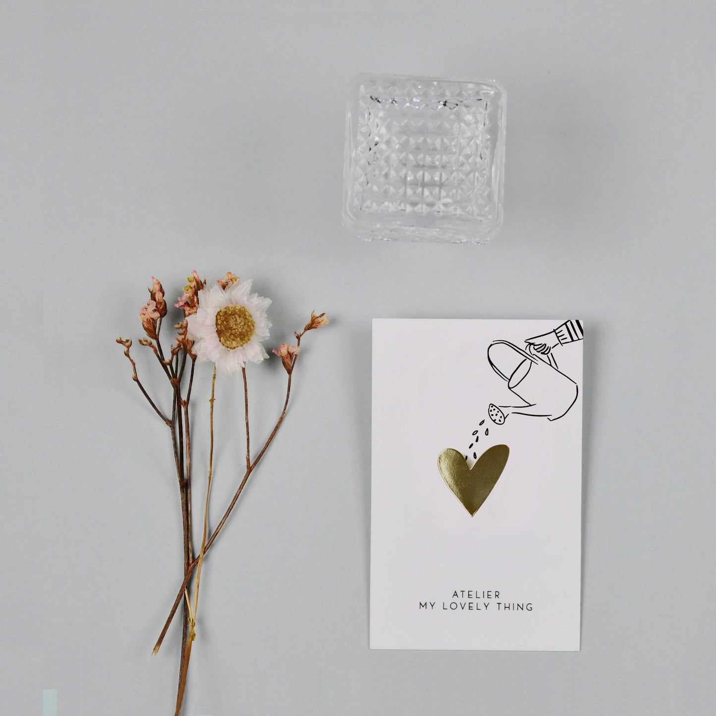 Card with heart illustration and text, accompanied by a dried flower and clear glass container on a light gray background.