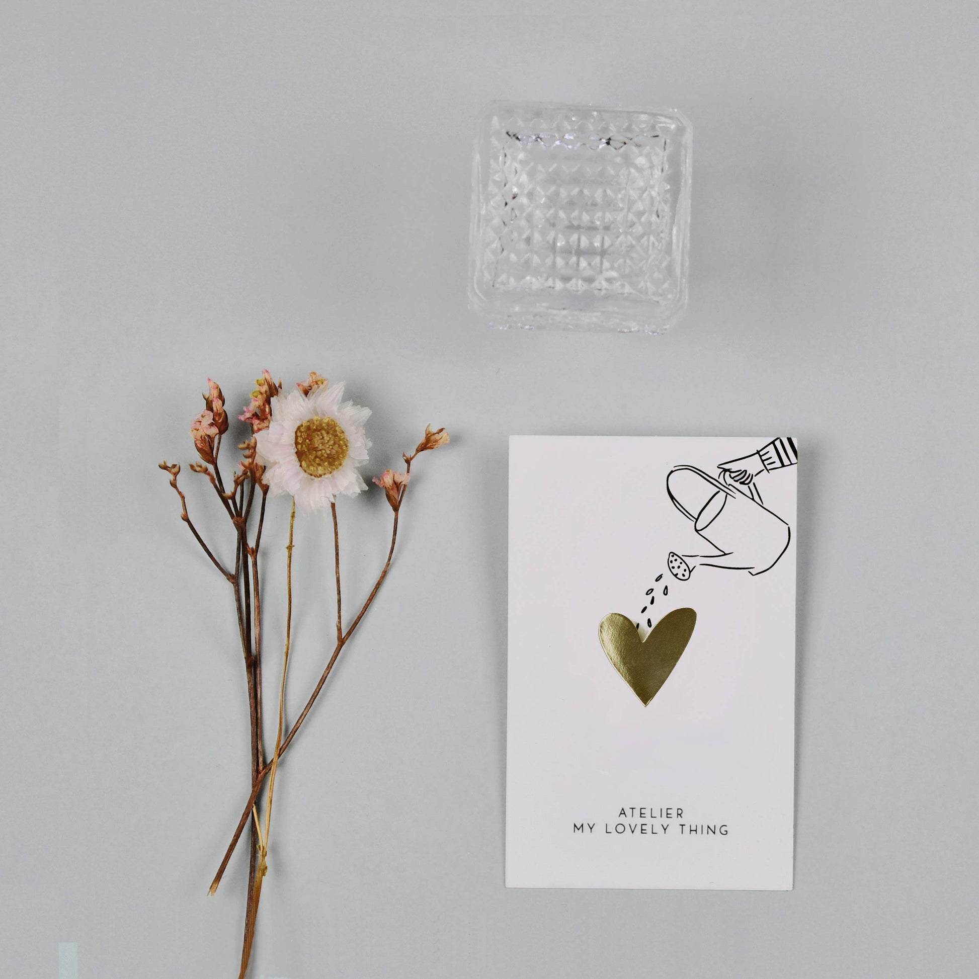 Card with heart illustration and text, accompanied by a dried flower and clear glass container on a light gray background.