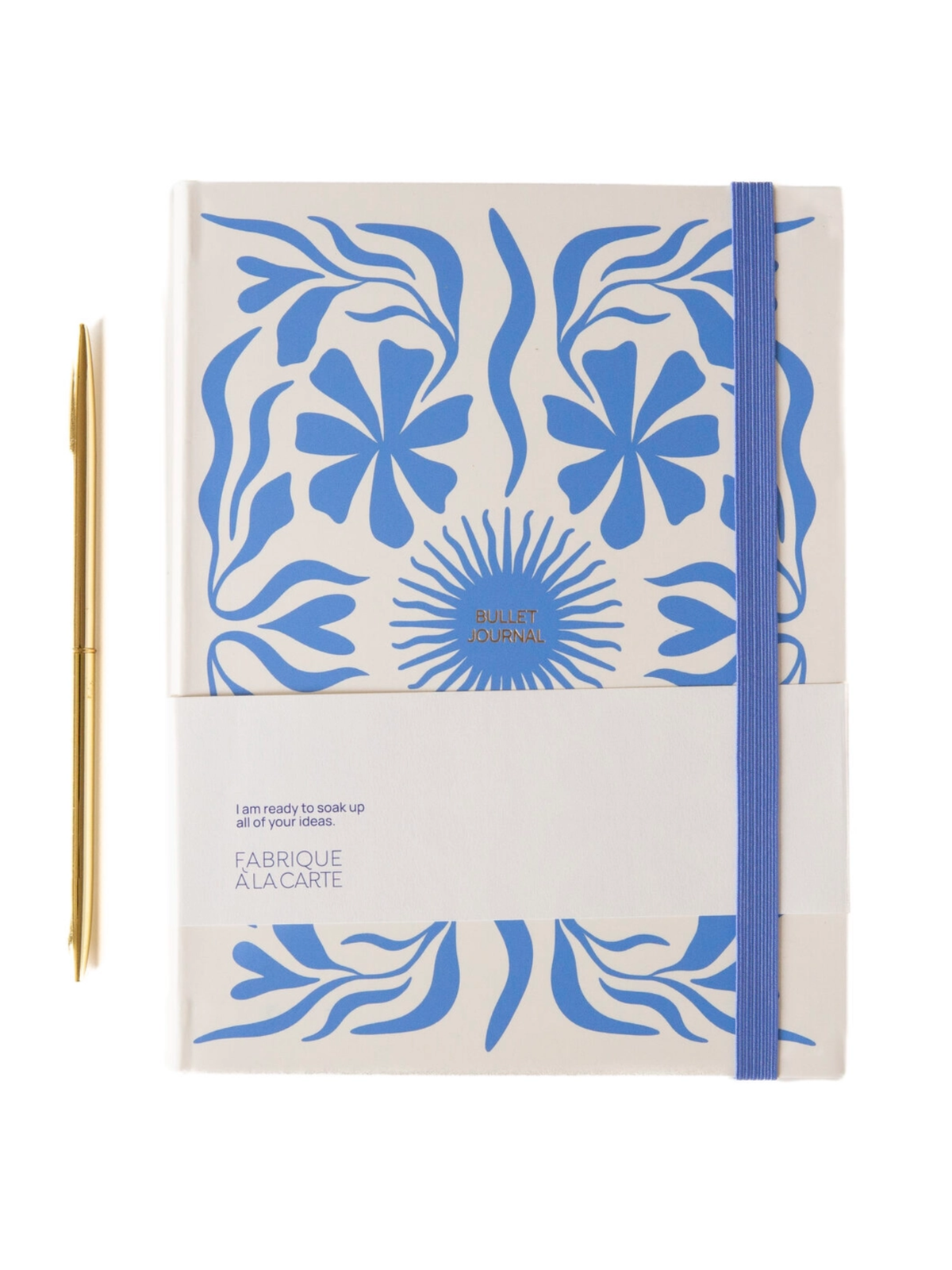 A Bullet Design notebook with a cream cover decorated with a blue floral pattern, held together by a blue elastic band, shown next to a gold pen on a white background.