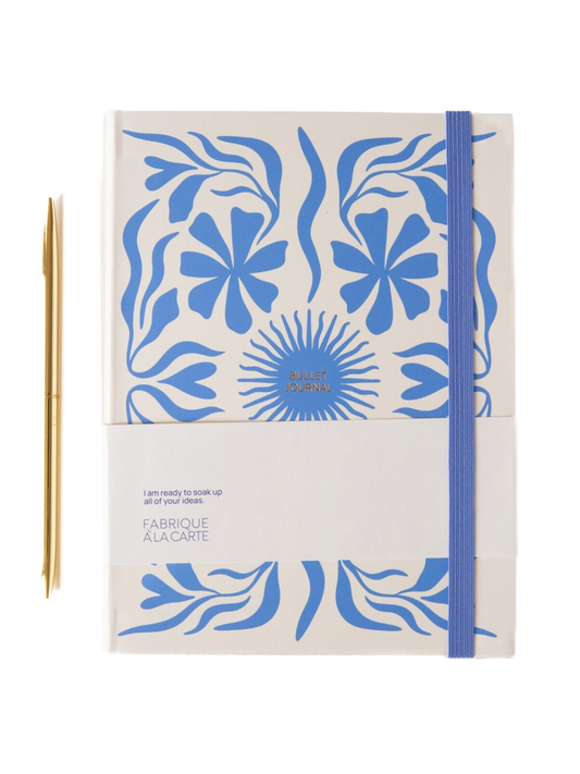A notebook with a cream-colored cover featuring a blue floral and leaf pattern, a blue elastic closure band, and a small orange bookmark tab at the bottom left. The cover includes the text 'I am ready to soak up all of your ideas.