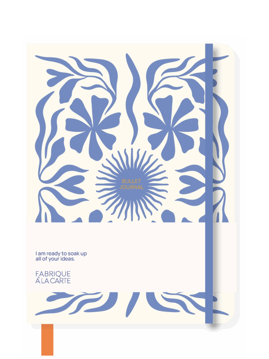 A notebook with a cream-colored cover featuring a blue floral and leaf pattern, a blue elastic closure band, and a small orange bookmark tab at the bottom left. The cover includes the text 'I am ready to soak up all of your ideas.