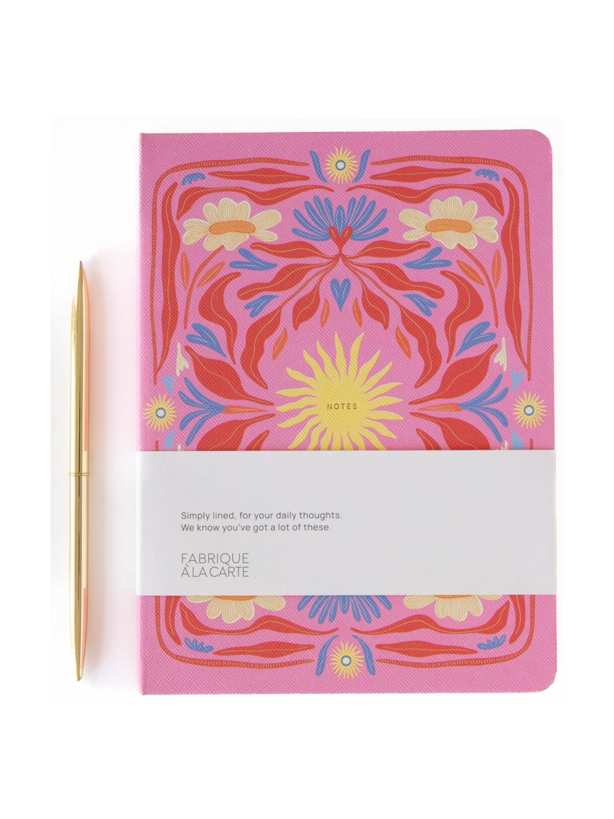 A pink Fabrique À La Carte notebook with a colorful floral mandala pattern, wrapped in a white paper band, shown next to a gold pen on a white background.