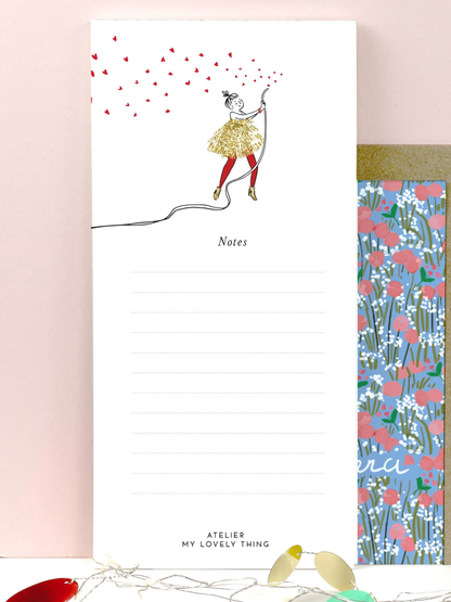 A white lined notepad with a whimsical illustration of a girl blowing red heart-shaped balloons, shown standing upright next to a floral-patterned notepad. The notepad has the text 'Notes' and 'ATELIER MY LOVELY THING' printed on it.