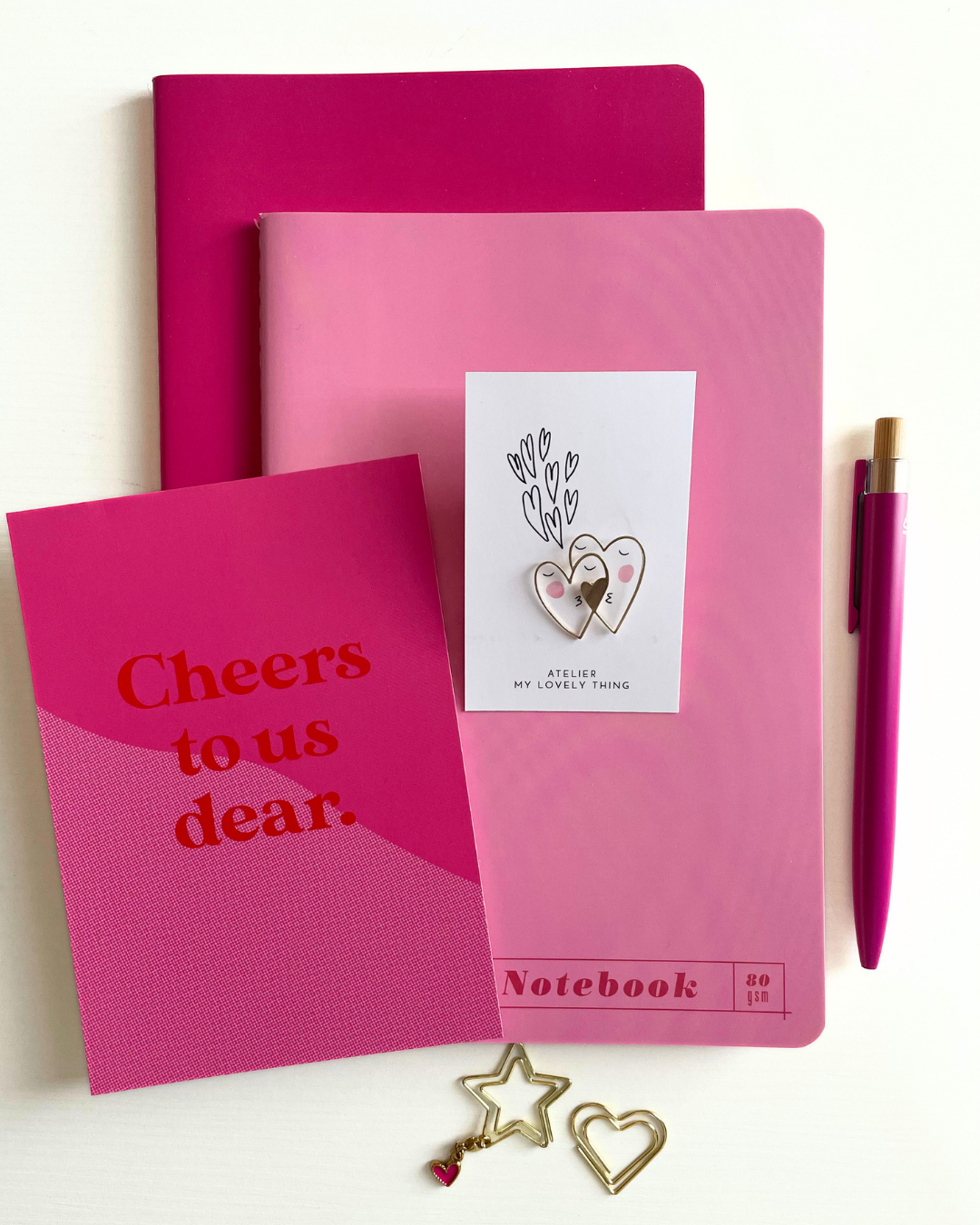 Pink notebook with 'Cheers to us dear.' text, a purple pen, and a small card on a white background.