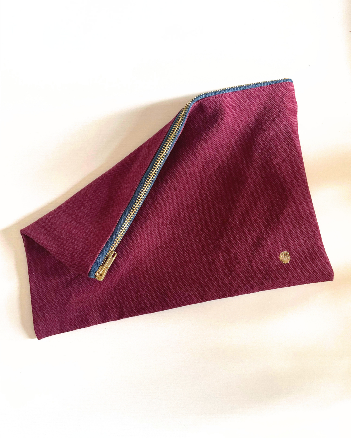 A deep burgundy felt pouch with a gold zipper and a small gold emblem, shown on a plain off-white background.