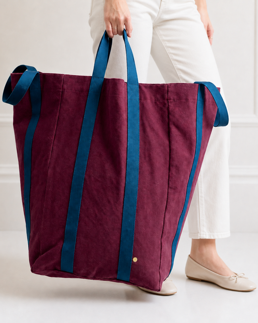A large canvas tote bag in burgundy with dark teal-blue handles and side trim, shown against a plain white background.