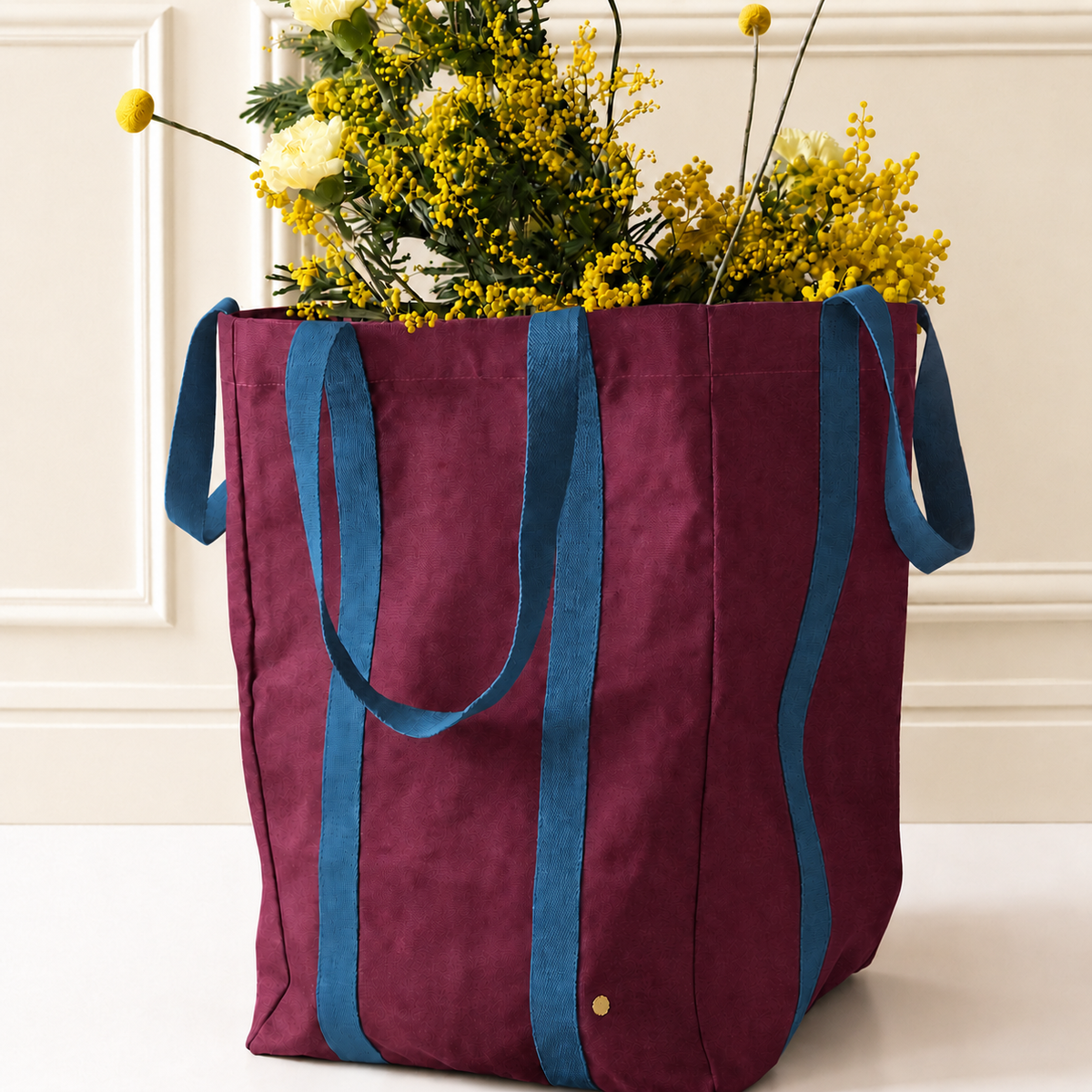 A large burgundy canvas tote bag with blue fabric handles and side straps, filled with yellow flowers, set against a cream paneled wall background.