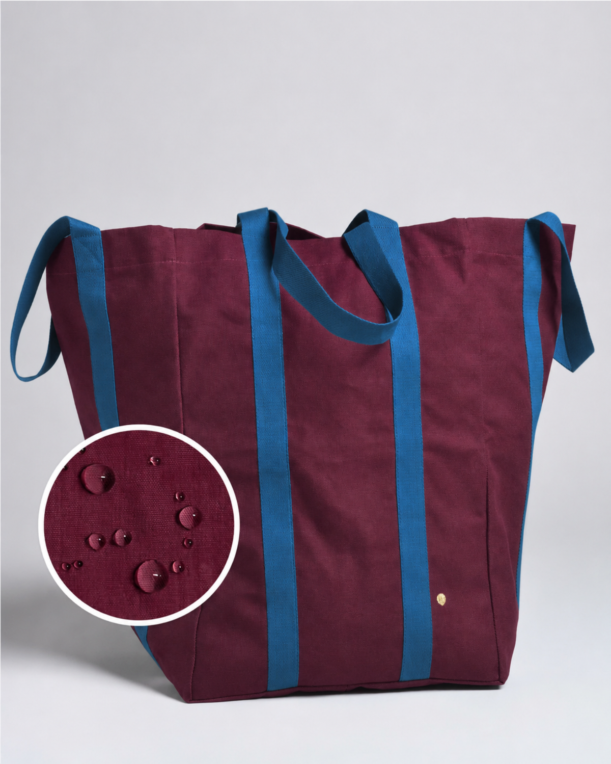 A large tote bag in deep burgundy with contrasting blue handles and side straps, shown on a light gray background with a circular inset detail of water droplets on the fabric.