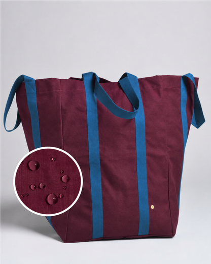 A large tote bag in deep burgundy with contrasting blue handles and side straps, shown on a light gray background with a circular inset detail of water droplets on the fabric.