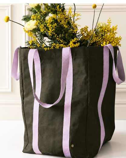 An olive green canvas tote bag with wide lavender-purple fabric handles, filled with yellow flowers, set against a white paneled wall background.