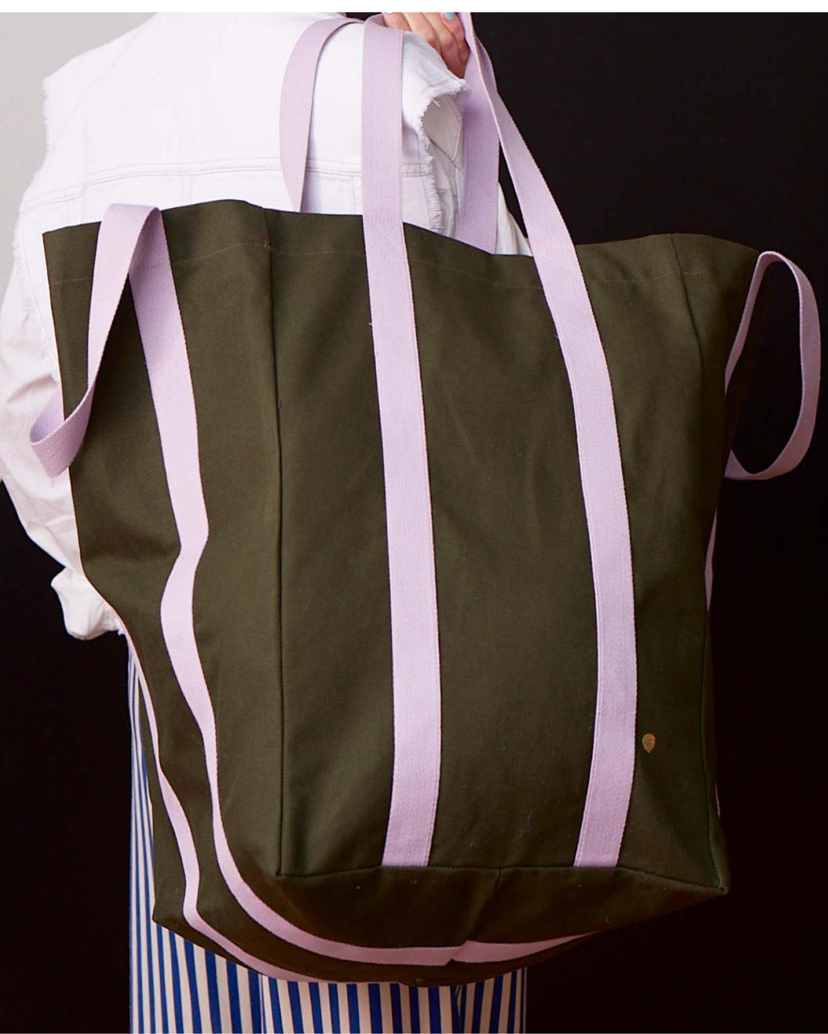 An olive green canvas tote bag with wide light purple-pink straps, carried over the shoulder of a person wearing a white jacket and blue-and-white striped skirt against a dark background.