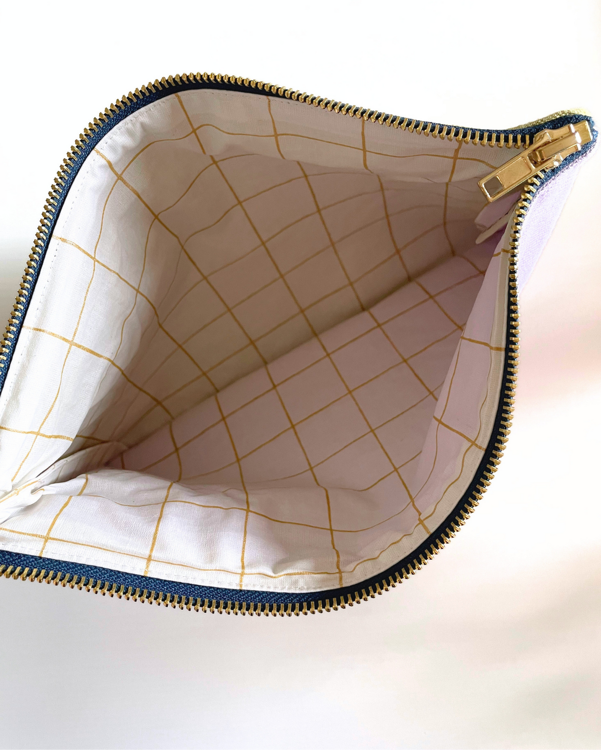 An open zippered pouch or cosmetic bag shown from above, revealing a white interior lined with a gold grid pattern. The exterior edge shows a navy blue trim and a gold zipper.