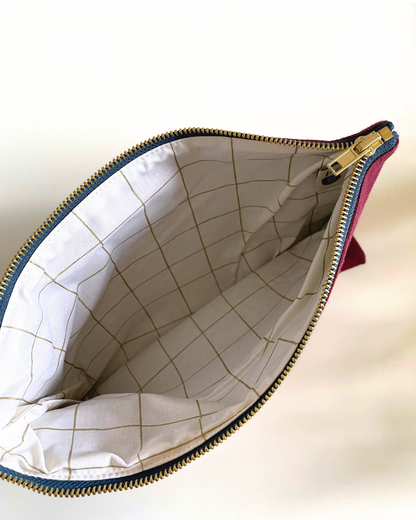 An open zippered pouch or clutch shown from above, revealing a cream-colored interior lined with a subtle gold grid pattern. The exterior fabric at the top edge appears magenta, and the zipper track is gold-toned.