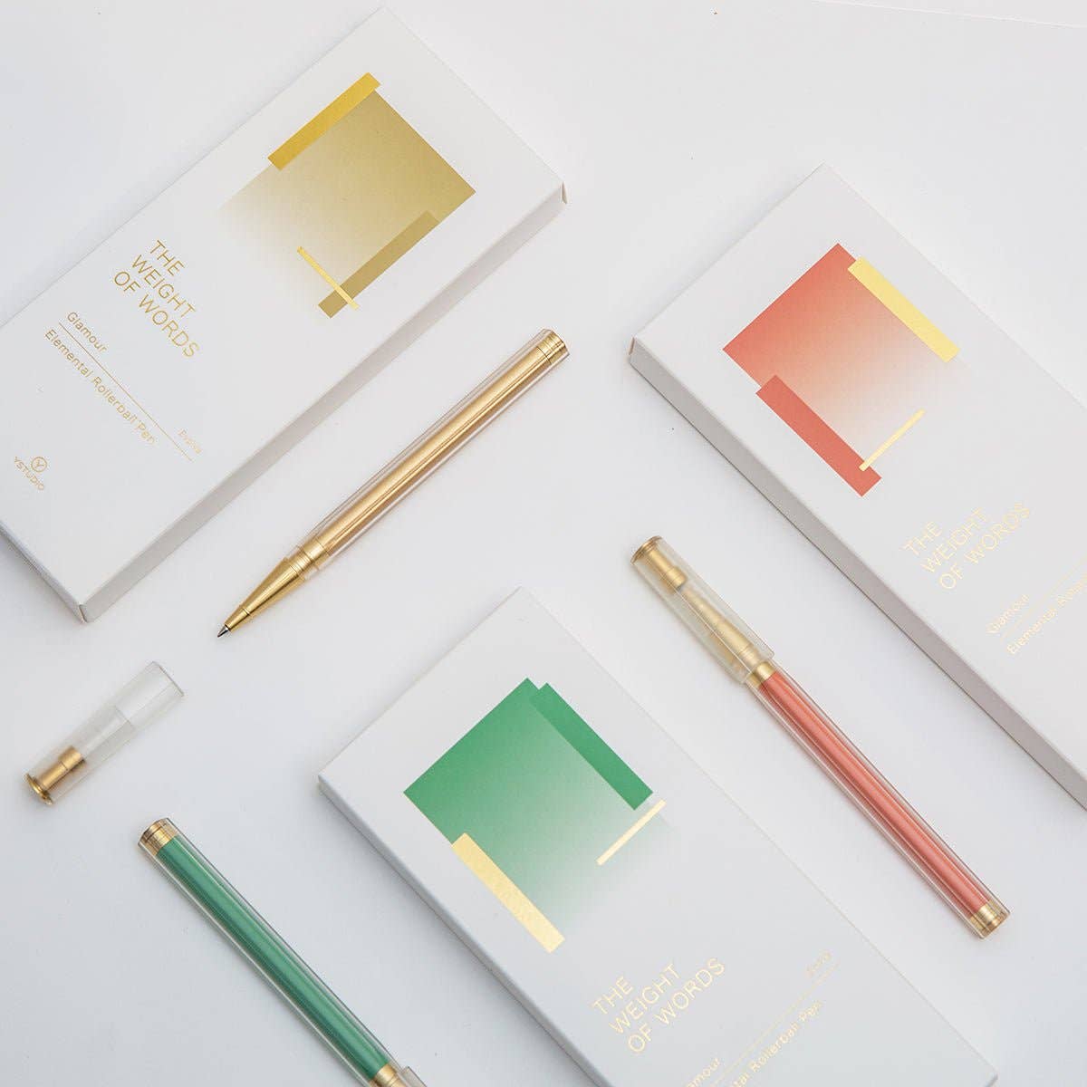 Three gold-finish ballpoint pens in green, pink, and gold are arranged on a white surface alongside their white packaging boxes, which feature minimalist geometric designs in green, pink, and gold.