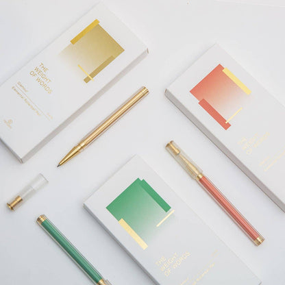 Three gold-finish ballpoint pens in green, pink, and gold are arranged on a white surface alongside their white packaging boxes, which feature minimalist geometric designs in green, pink, and gold.