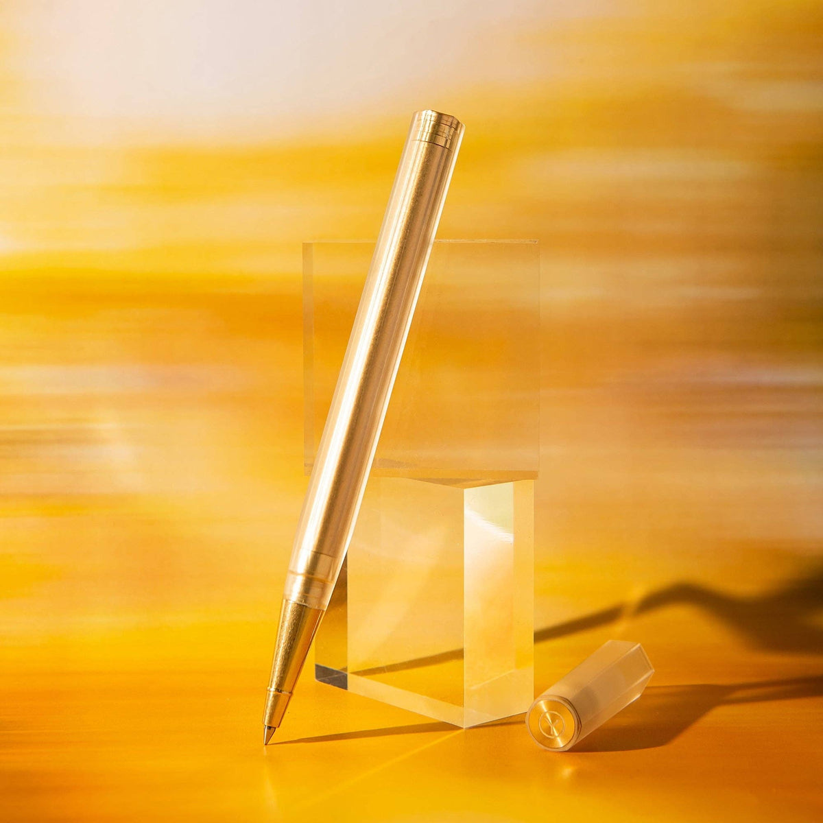 A metallic gold pen displayed upright on a clear acrylic block, with its cap placed beside it on a warm golden-yellow background.