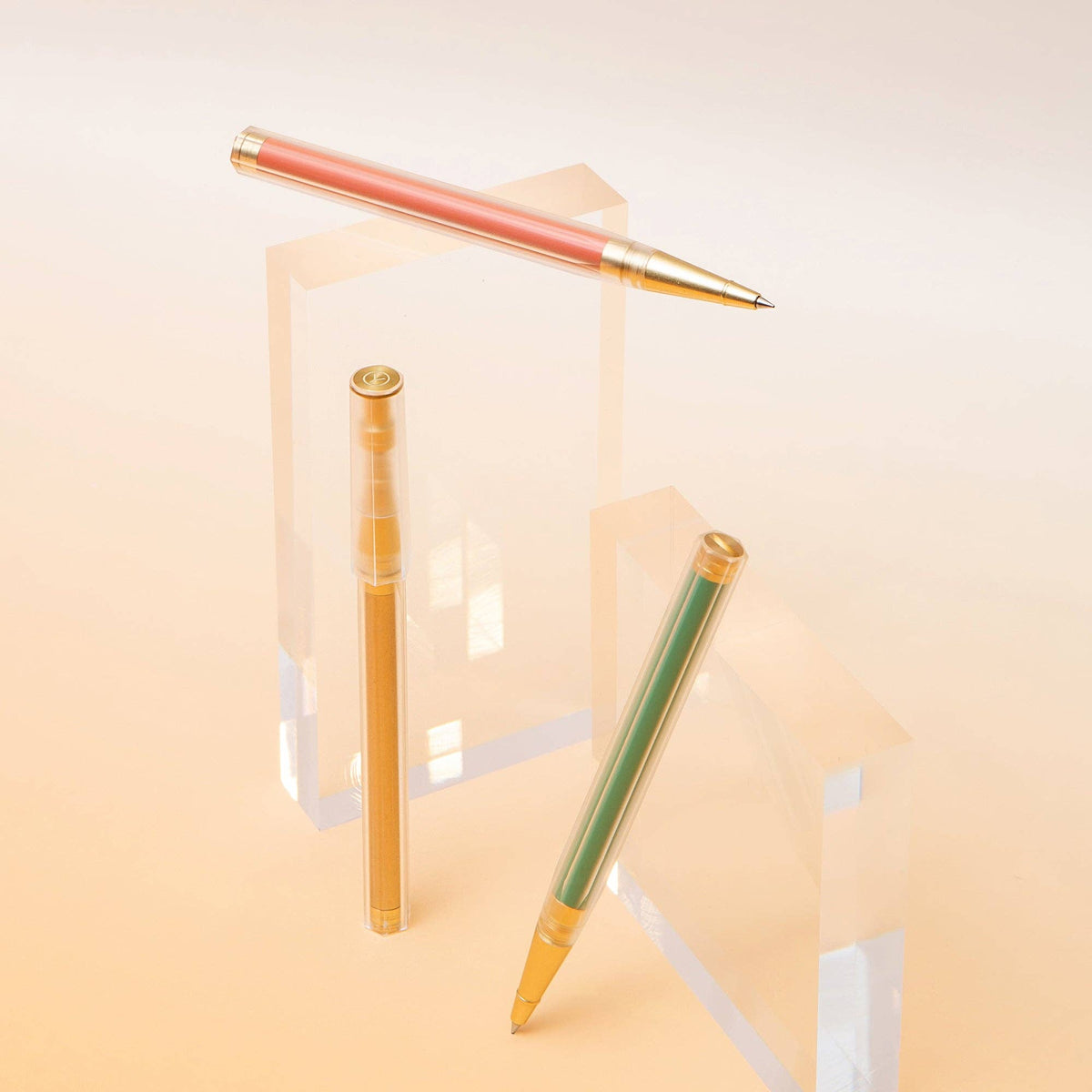 Three slim retractable pens displayed on clear acrylic blocks against a soft warm beige background. One pen is horizontal at the top, one is vertical in the center, and one is diagonal at the bottom right.