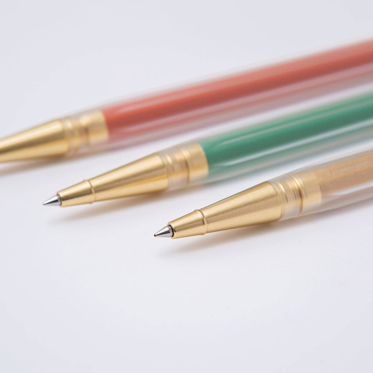 Three retractable ballpoint pens with gold metal clickers and tips, shown diagonally on a clean white background. The pen barrels are translucent, revealing coral red, green, and beige ink colors.