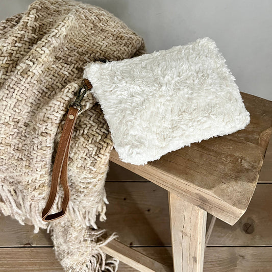 A fluffy off-white faux-shearling clutch bag with a brown leather wristlet strap, resting on a rustic wooden stool next to a woven beige throw blanket.