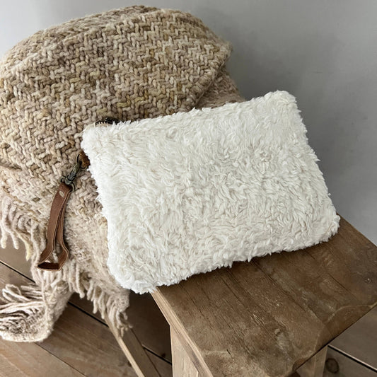 A fluffy off-white faux-shearling clutch bag with a brown leather wristlet strap, resting on a rustic wooden stool next to a woven beige throw blanket.