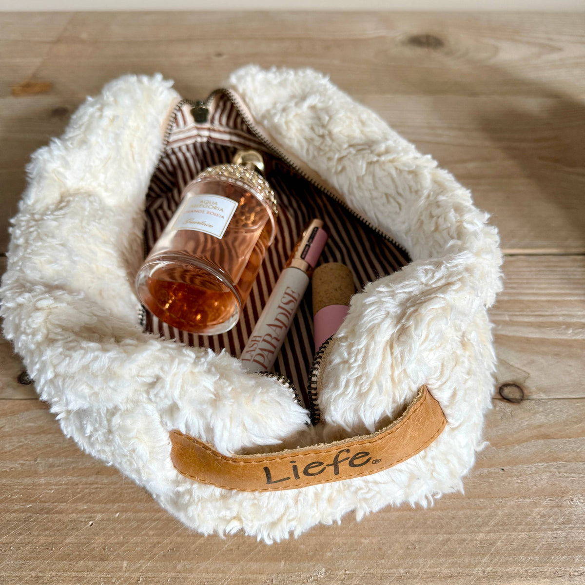 A fluffy cream faux-fur zip pouch with a tan leather handle embroidered with the word 'Liebe' in black script, shown open on a light wood surface. Inside are several cosmetic items, including a perfume bottle and lip product.
