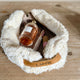 A fluffy cream faux-fur zip pouch with a tan leather handle embroidered with the word 'Liebe' in black script, shown open on a light wood surface. Inside are several cosmetic items, including a perfume bottle and lip product.