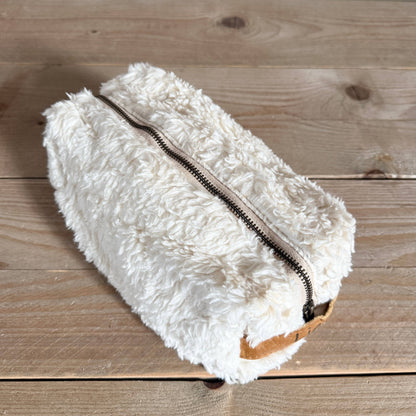 A fluffy off-white faux fur zippered pouch with a light brown leather accent tab, resting on a light brown wooden plank surface.