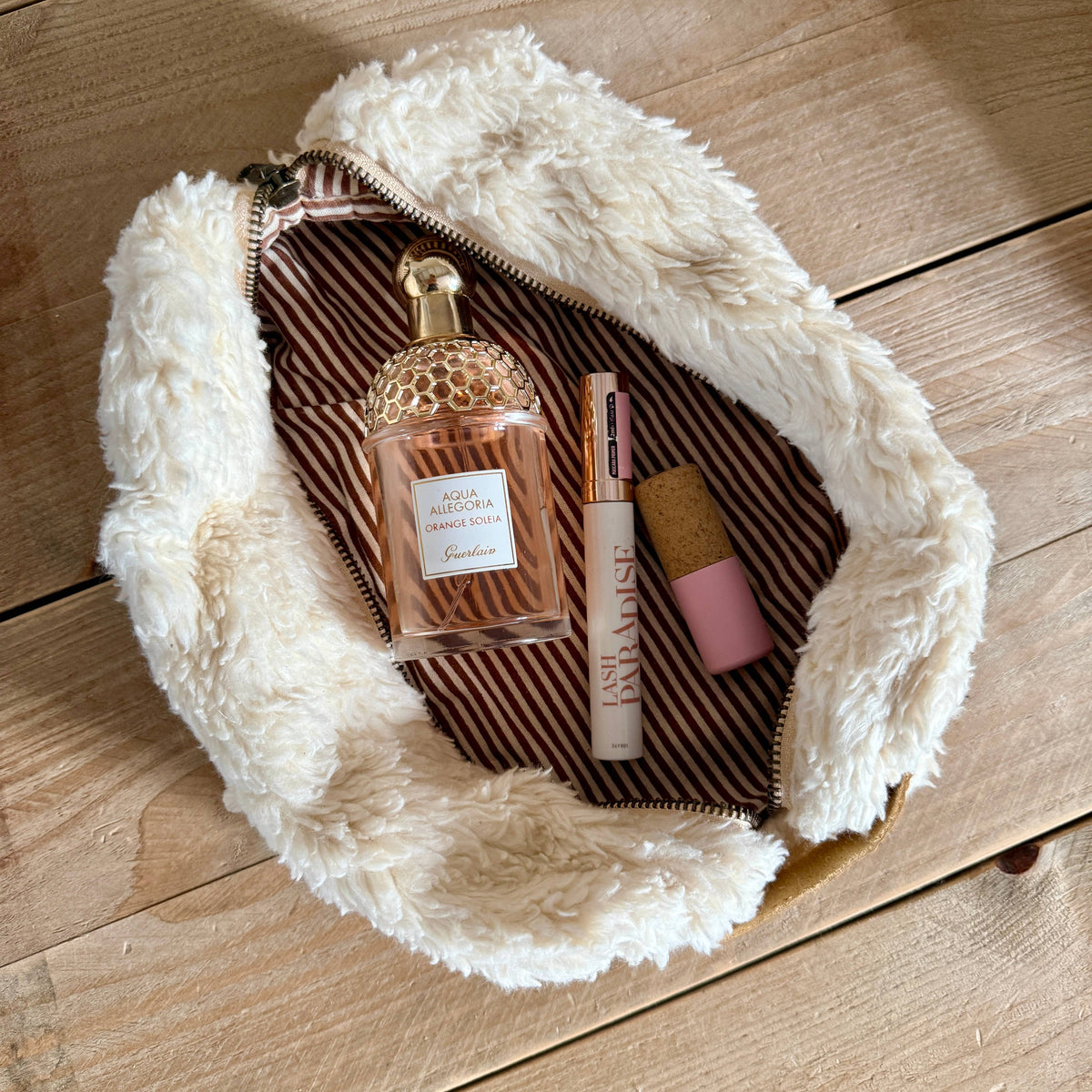 A fluffy cream faux-fur zip pouch with a brown and white striped interior, shown open on a light wood surface with perfume, mascara, and a pink lip product inside.