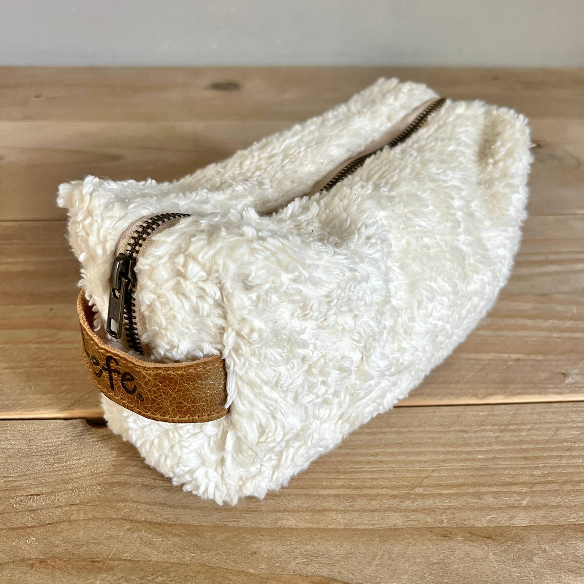 A fluffy off-white sheepskin or faux-shearling zip pouch with a brown leather tab and dark zipper, resting on a light wood tabletop.