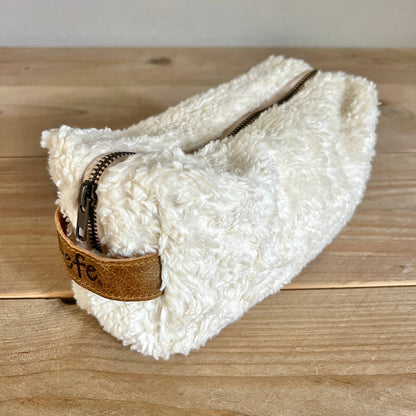 A fluffy off-white sheepskin or faux-shearling zip pouch with a brown leather tab and dark zipper, resting on a light wood tabletop.