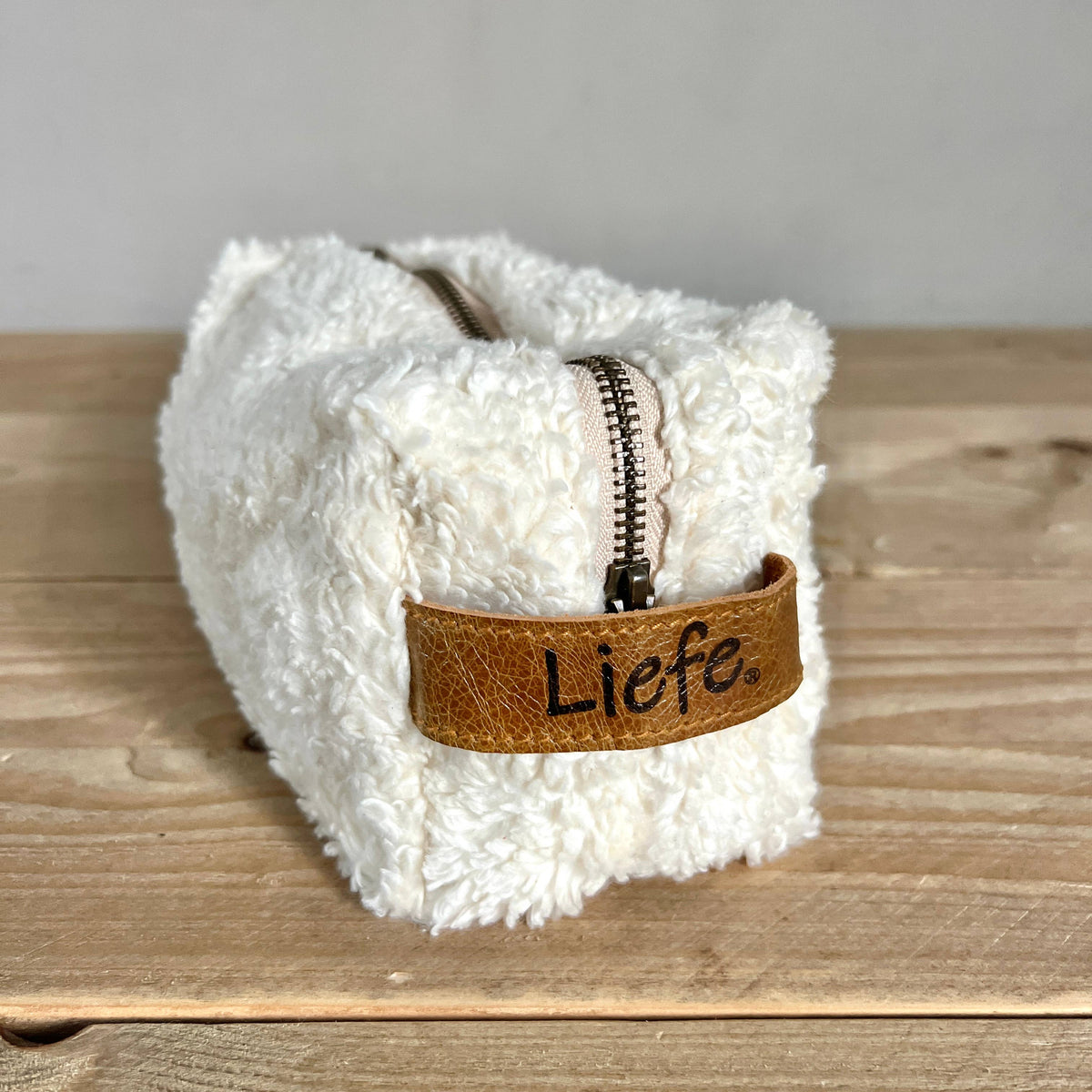 A white sherpa zippered pouch with a brown faux-leather tab that reads 'Liefе.' in black script, placed on a wooden surface against a neutral gray background.