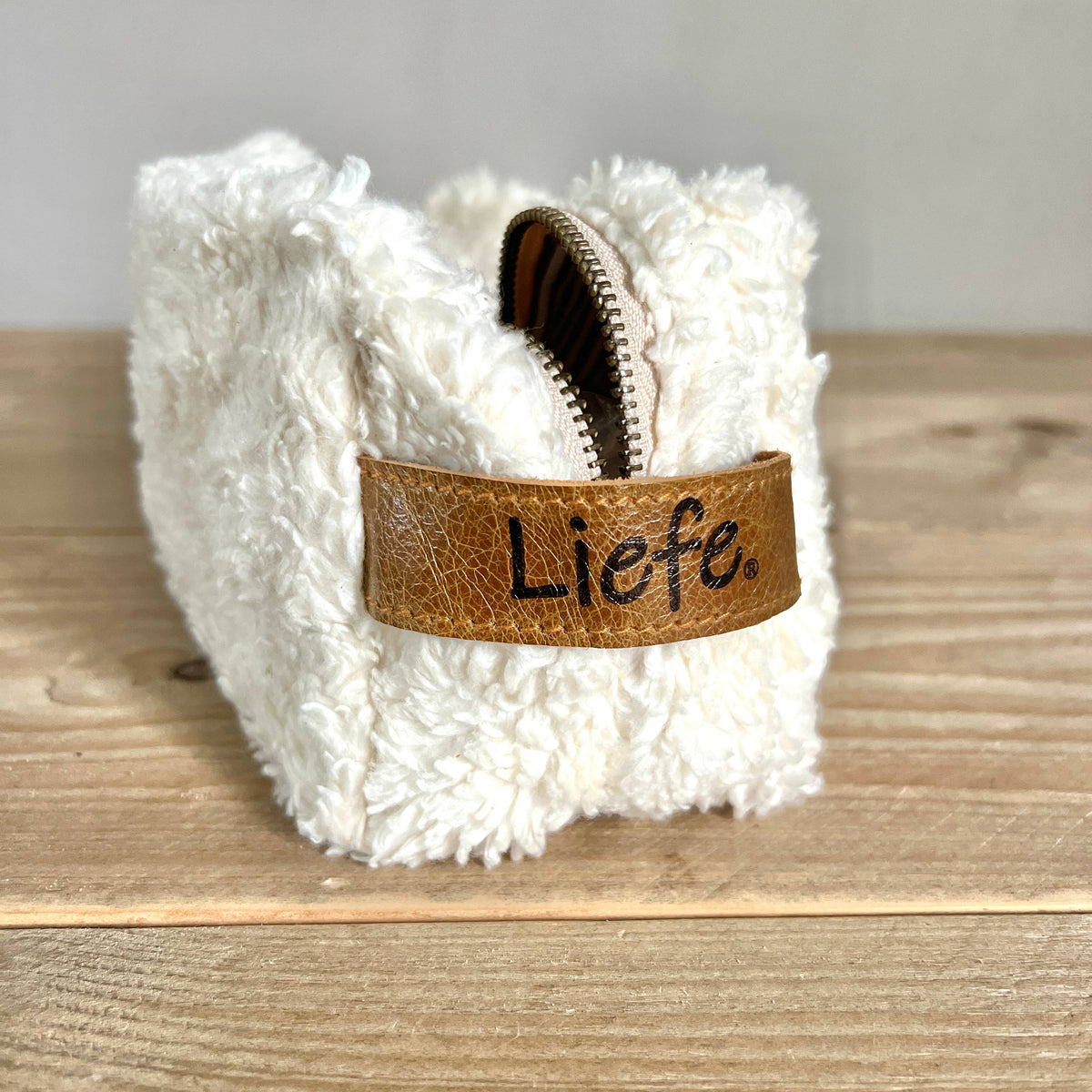 A small white faux-fur zip pouch with a brown leather strap across the front that reads 'Liefe.' in black script, photographed on a wooden surface against a light neutral background.