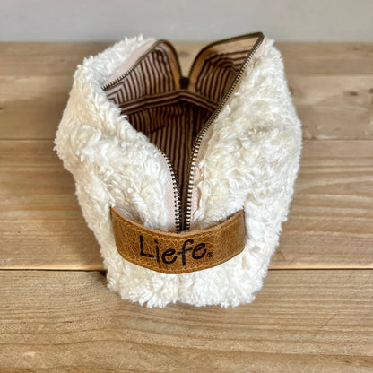 A white sherpa zippered pouch with a brown faux-leather strap featuring the embroidered brand name 'Liefe'.