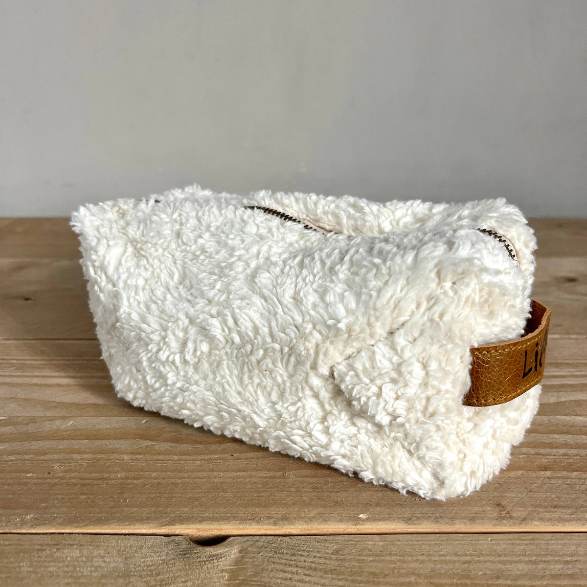 A white sherpa zippered pouch with a brown leather accent tab on the side, resting on a wooden tabletop against a neutral gray wall.