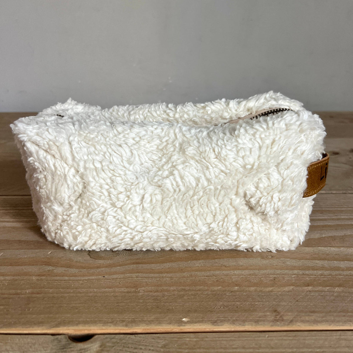 A white sherpa fleece zippered pouch with a small brown leather accent tab, resting on a rustic wooden surface against a neutral gray wall.