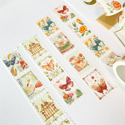 Several decorative washi tapes with vintage-style illustrated stamps are arranged on a white textured surface. The tapes feature butterflies, flowers, birds, and small architectural motifs in soft pastel colors, with one tape showing the word “PARIS” and another showing a birdhouse illustration.