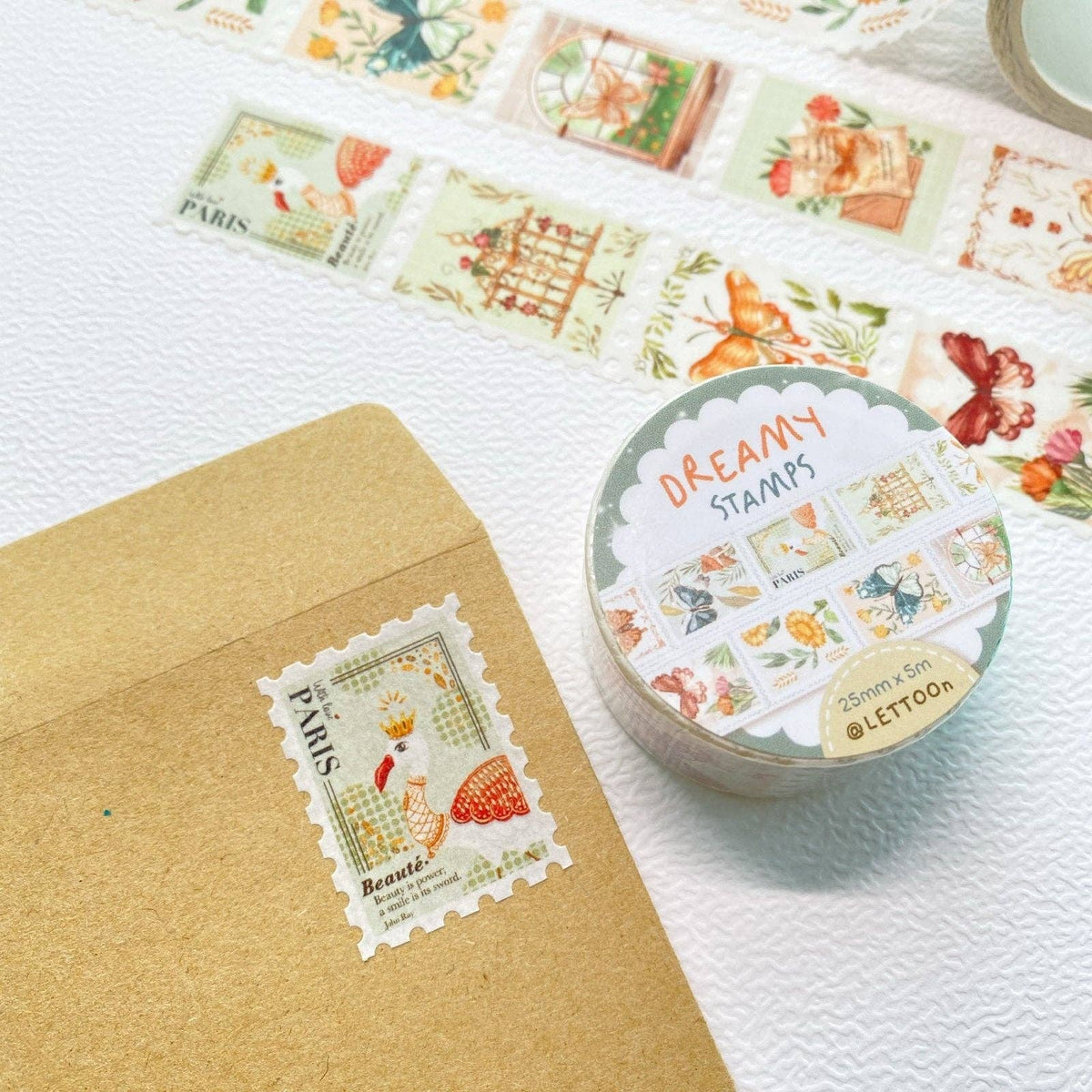 A round tin of decorative washi tape labeled 'DREAMY STAMPS' sits on a white textured surface beside a kraft envelope with a Paris-themed stamp design. Several illustrated stamp strips are arranged in the background.