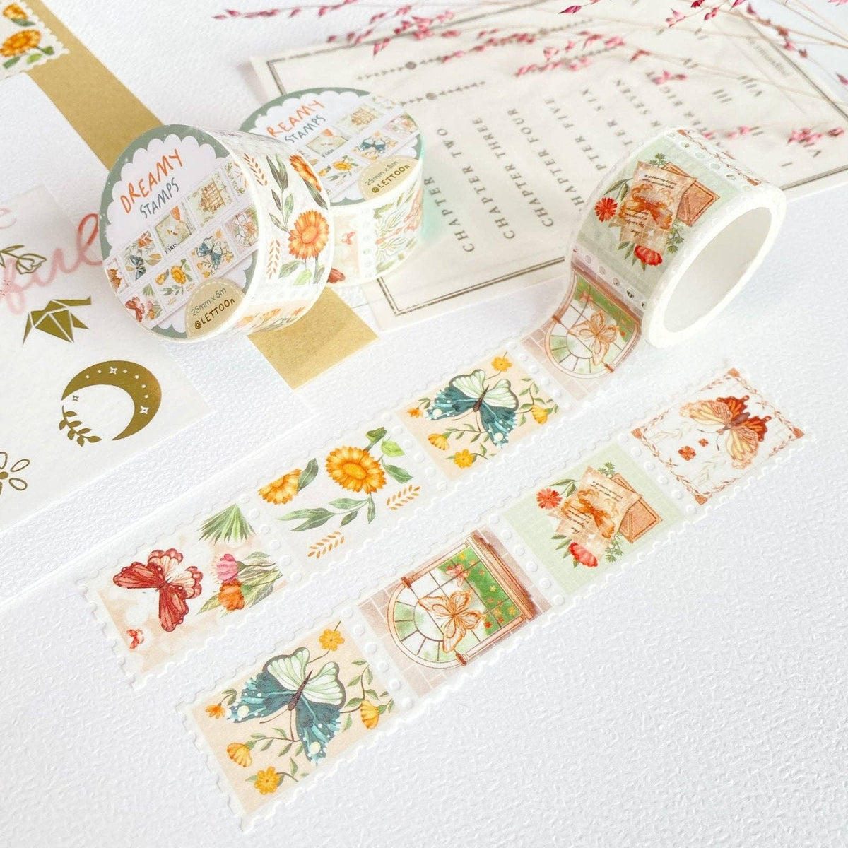 Two rolls of decorative washi tape with vintage-style illustrated stamps and floral motifs, displayed on a white textured surface with sample strips unrolled.