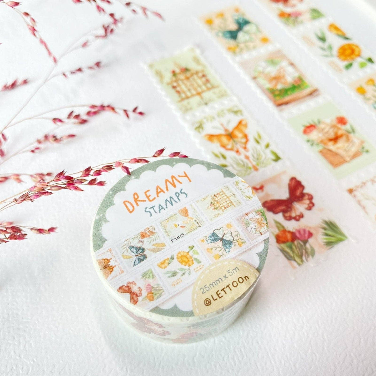 A roll of decorative washi tape labeled 'DREAMY STAMPS' by @LETOON, shown on a white textured surface with dried red flowers and other sample strips laid out behind it.
