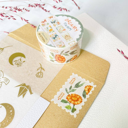 A roll of decorative washi tape labeled 'DREAMY STAMPS' sits on top of kraft paper envelopes and a white card with gold foil designs. The tape has a cream background with small illustrated stamps in orange, yellow, green, and blue, and the roll itself is white with a green border.