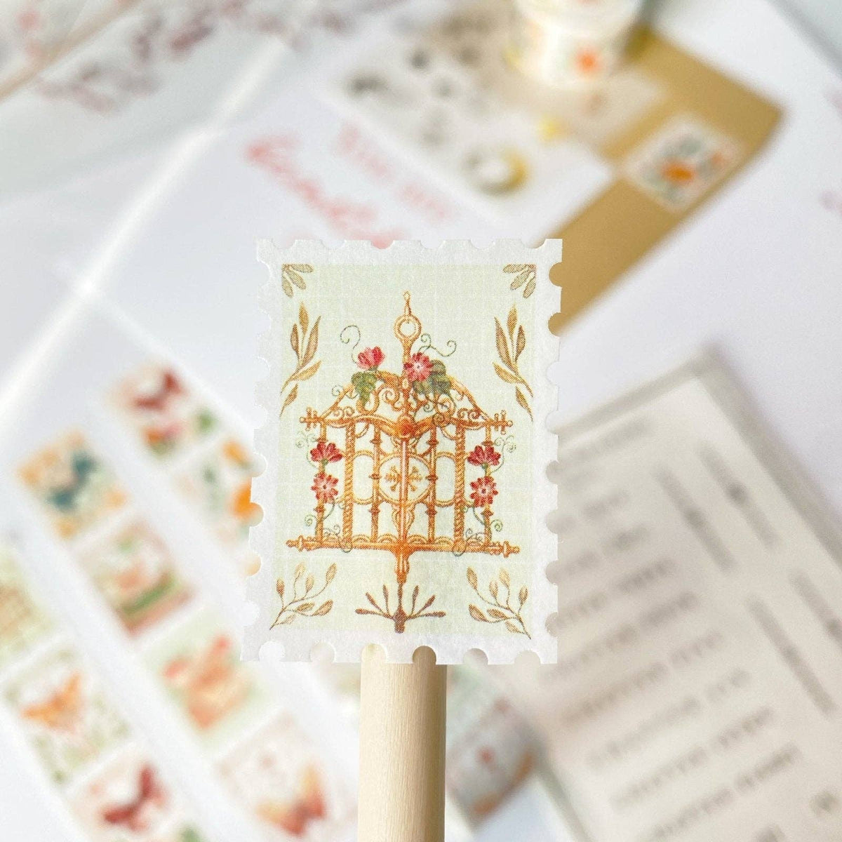A close-up of a decorative postage-stamp-style sticker or decal mounted on a wooden stick, featuring an ornate golden gate with red flowers and green leaves on a cream background. Other similar decorative pieces are softly blurred in the background.