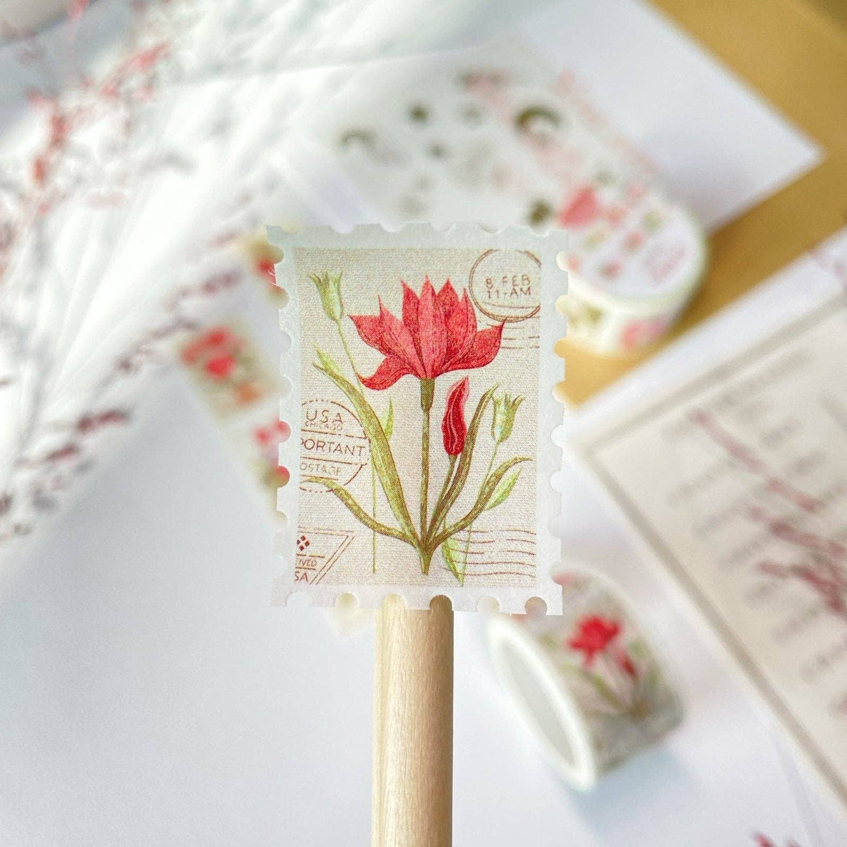 A close-up of a rectangular paper stamp or label with a red tulip illustration and vintage-style postmark text, mounted on a wooden stick. The background is softly blurred with similar floral paper items and a light tabletop surface.