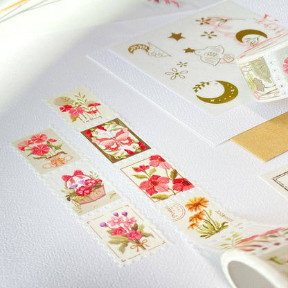 A roll of decorative washi tape with vintage-style floral and celestial illustrations is unrolled on a white surface, alongside matching decorative paper sheets and a gold envelope.